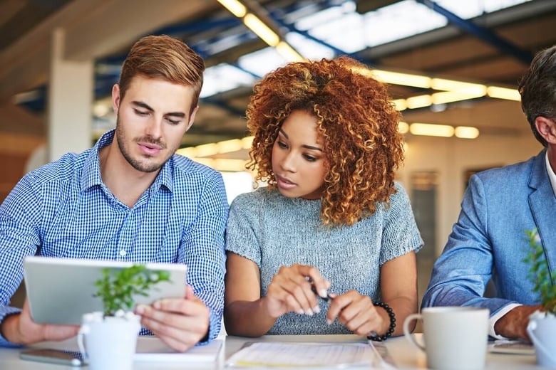 Two coworkers sit at a conference table and look at a tablet together