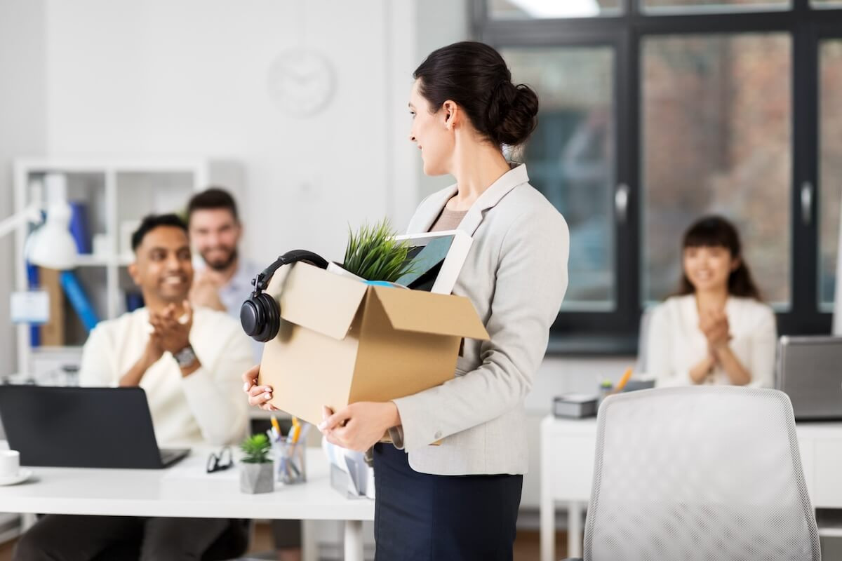 Managing employee absenteeism: employee looking back at her colleagues while carrying a box filled with her belongings