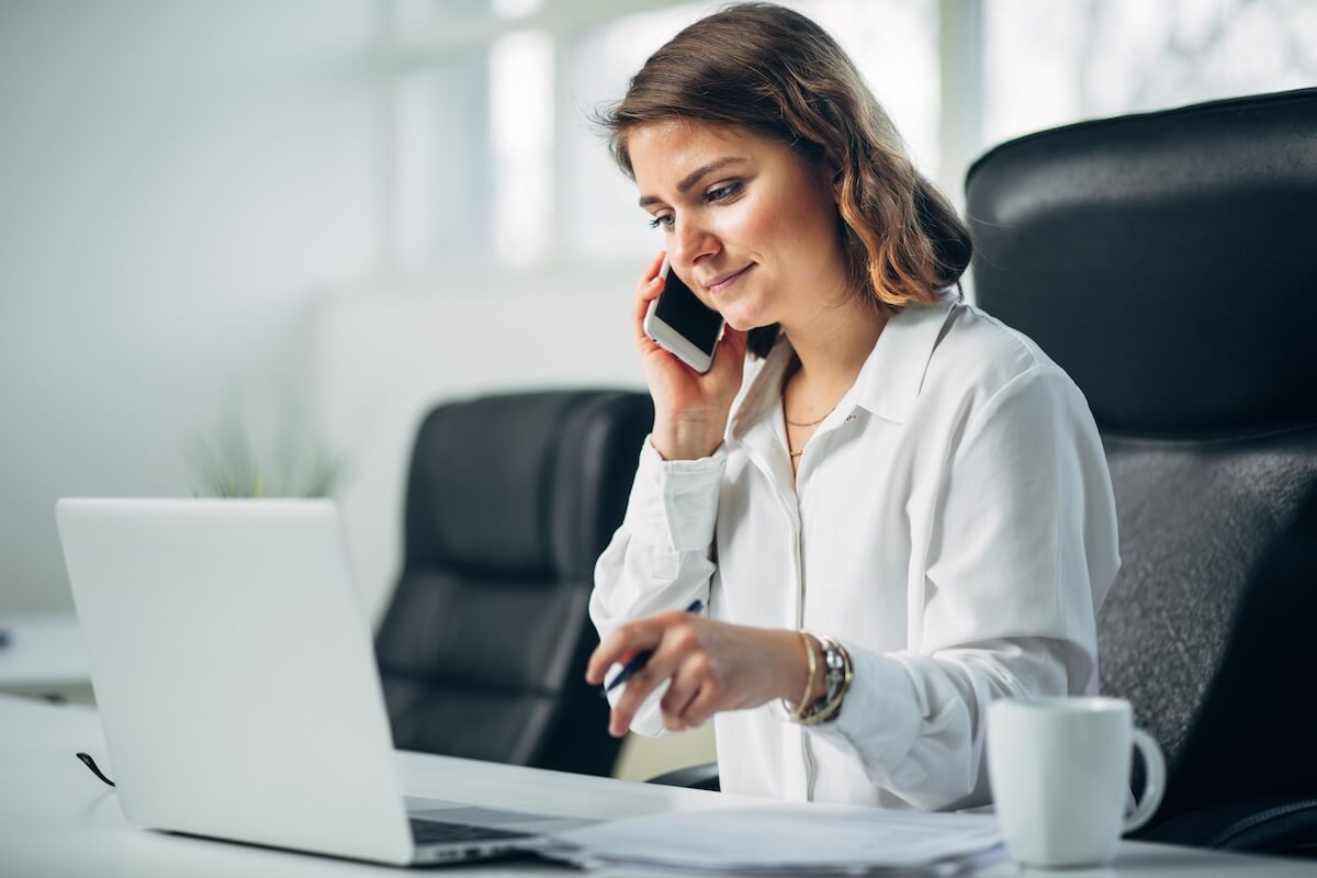 A woman sits in front of a laptop and talks on a cellpho